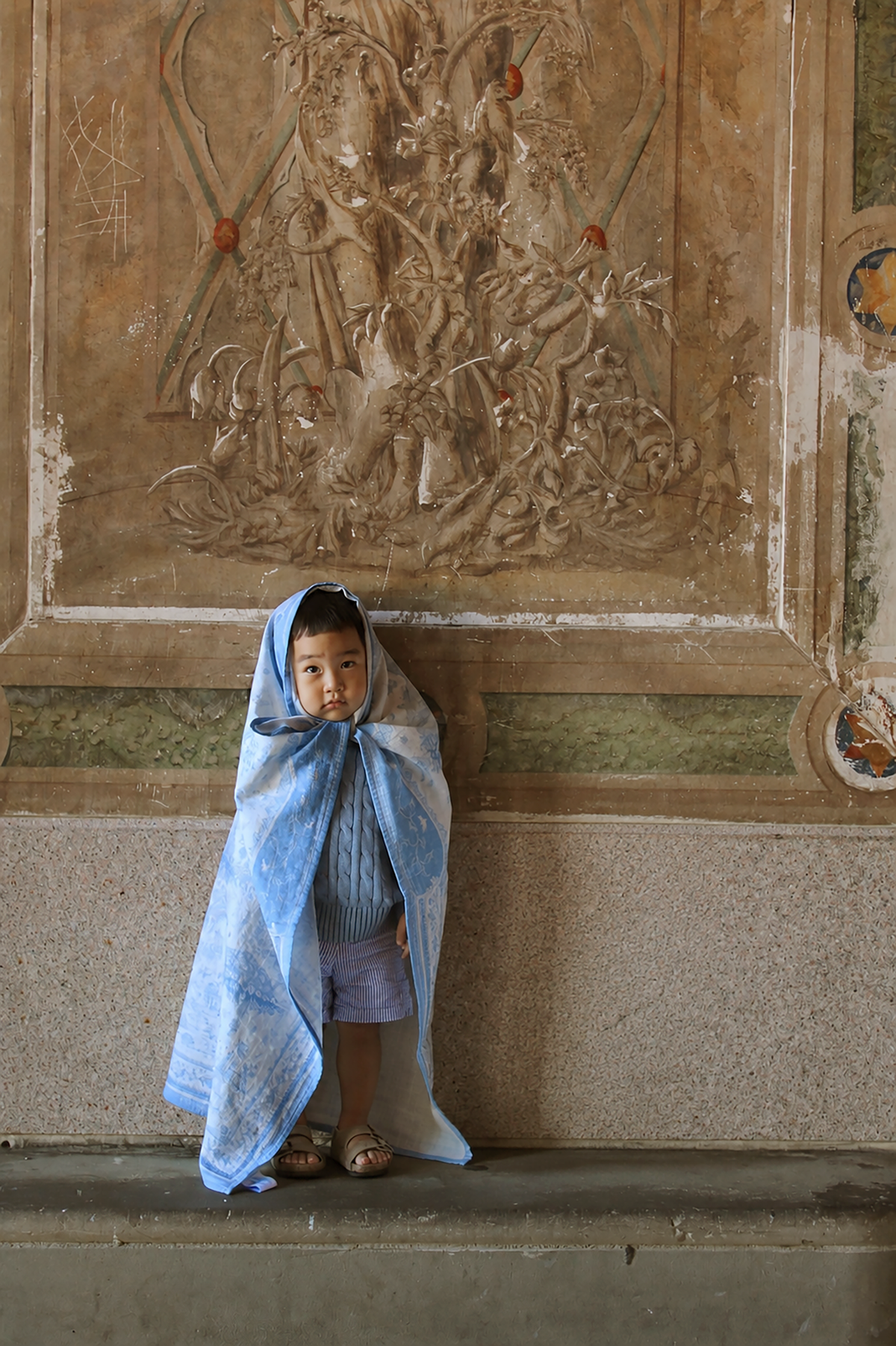 A young child stands on a stone ledge in front of a large carved wall panel, wrapped in a light blue and white patterned cloth.