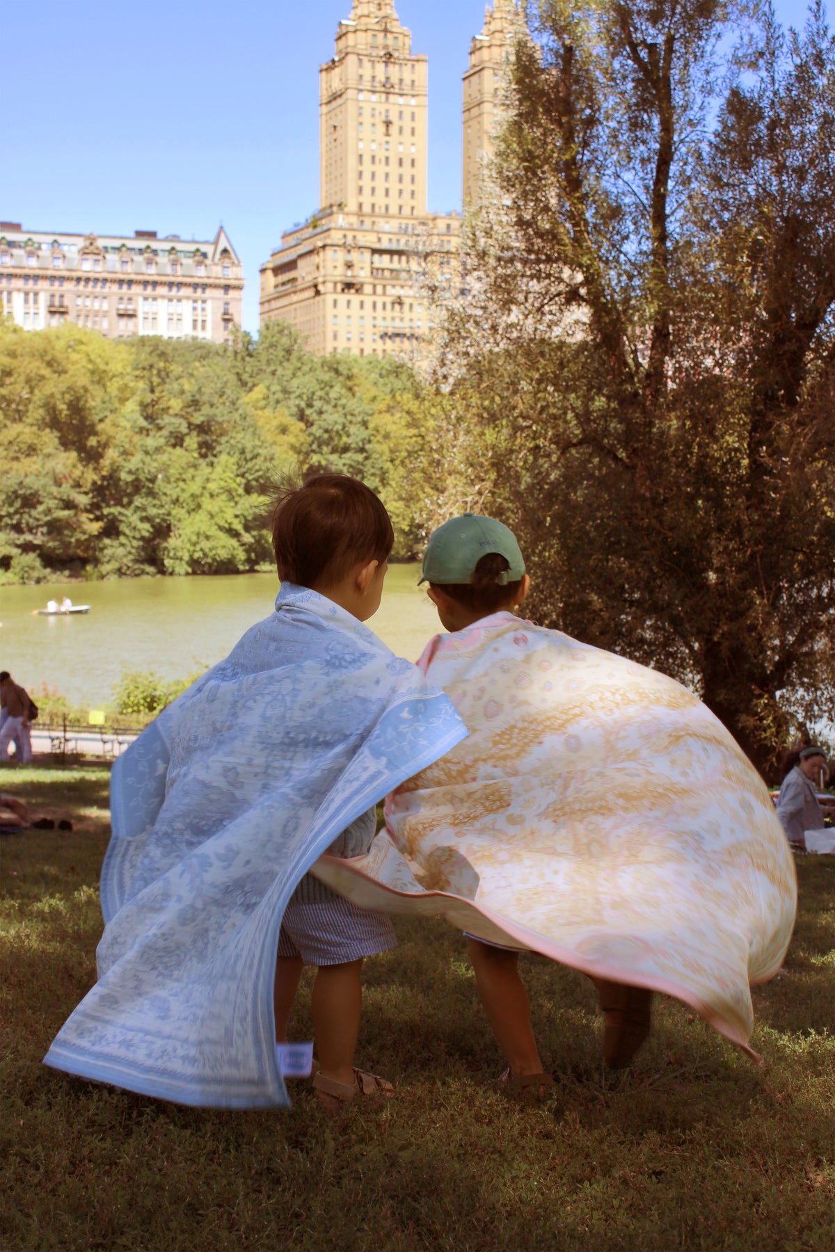 Two children standing on grass in a park, wrapped in patterned blankets, with a lake, trees, and tall buildings in the background.